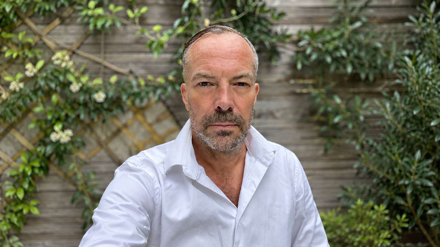 Portrait of filmmaker Simon Rumley wearing a white shirt against a garden backdrop.