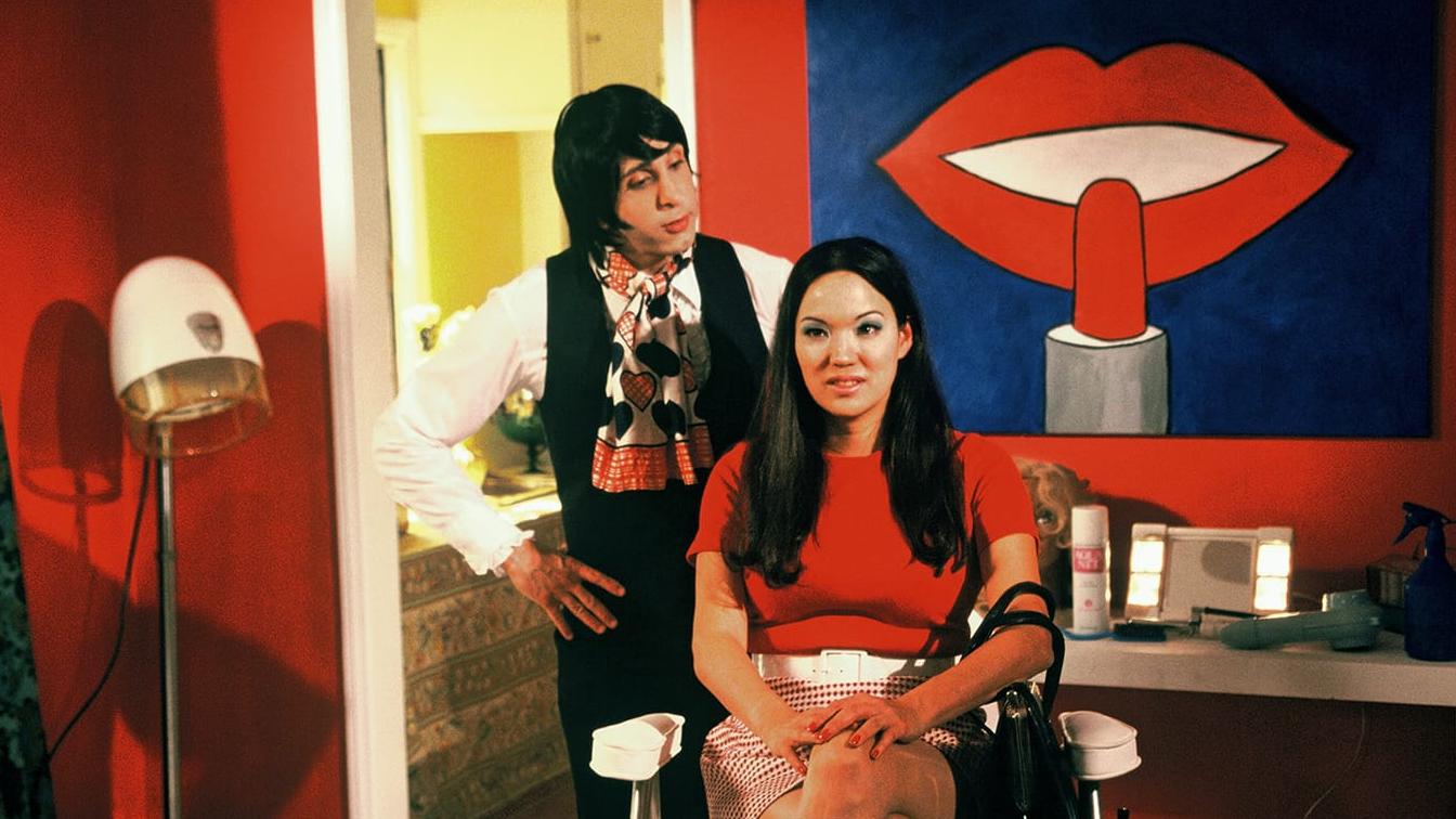 Anna Biller as Barbi in Viva, seated in a bright red salon beneath a painting of lips.