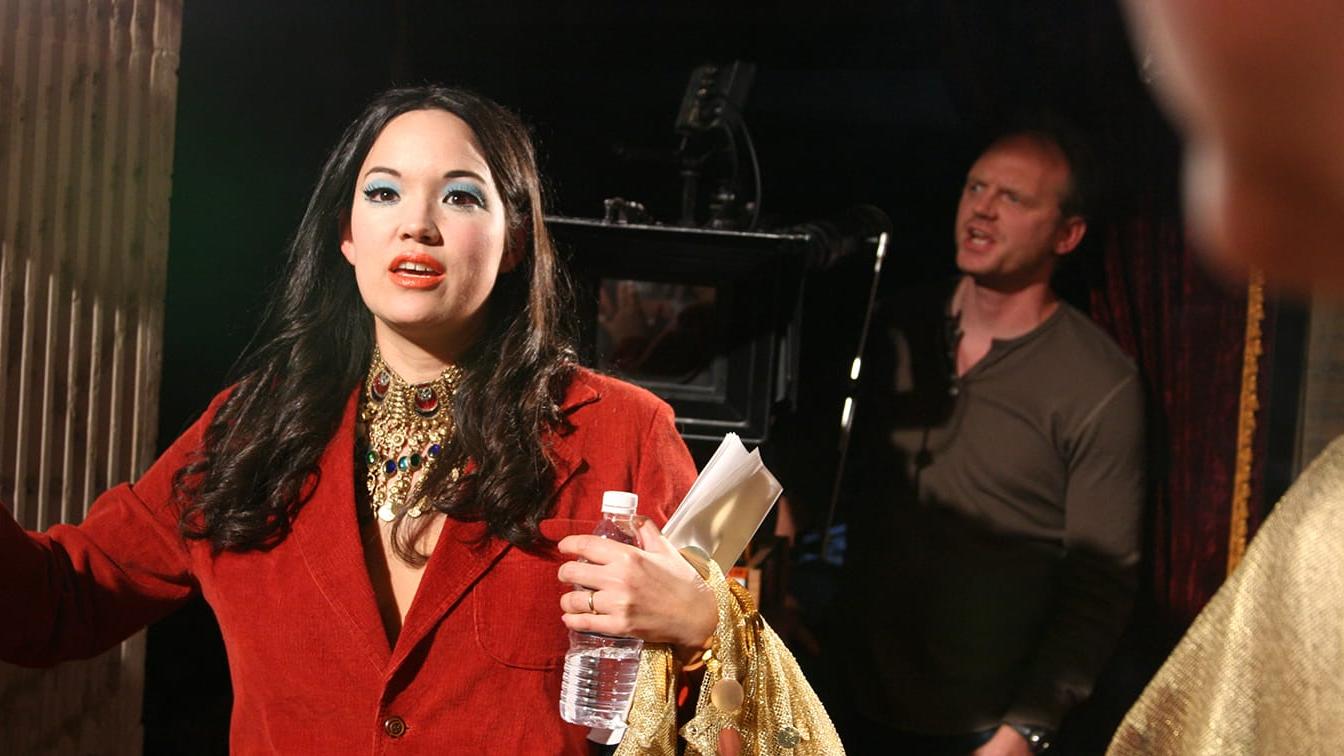 Anna Biller on the Viva set in a red jacket, with film equipment visible behind her.